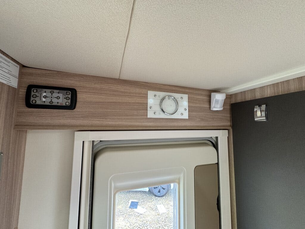 A portion of a room inside the 2018 Burstner Ixeo TL680 G features a wooden panel wall with a small circular clock, a remote control holder, and a corner sensor. Below is a partially open door with a black frame and small window, revealing the floor outside.