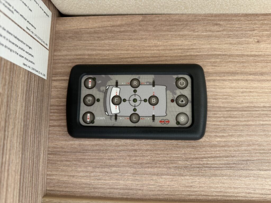 A black control panel with various buttons and a display is mounted on a wooden surface inside the 2018 Burstner Ixeo TL680 G. The buttons are marked with symbols and letters, including "F," "R," "B," "S," and "UP" and "DOWN" arrows.