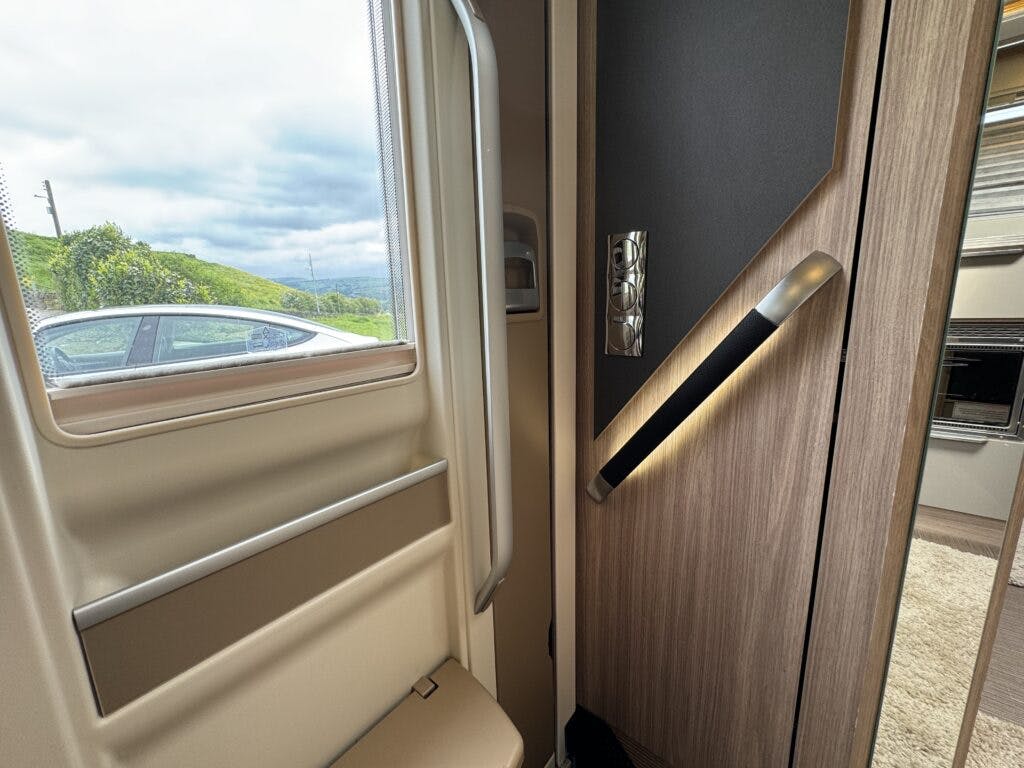 Interior of a modern 2018 Burstner Ixeo TL680 G showing a doorway and built-in seating. The door is partially open, revealing an outdoor view with green hills and a cloudy sky. The interior features wooden finishes, and a car is visible outside through the window.