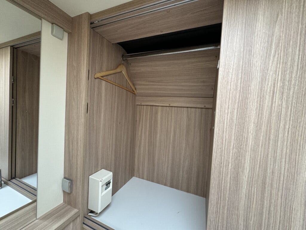 The image shows an empty wooden wardrobe with a white base, a single beige clothes hanger, and a small white safe inside. The wardrobe is part of a light wood-themed room within the 2018 Burstner Ixeo TL680 G and has a large mirror reflecting part of the interior.