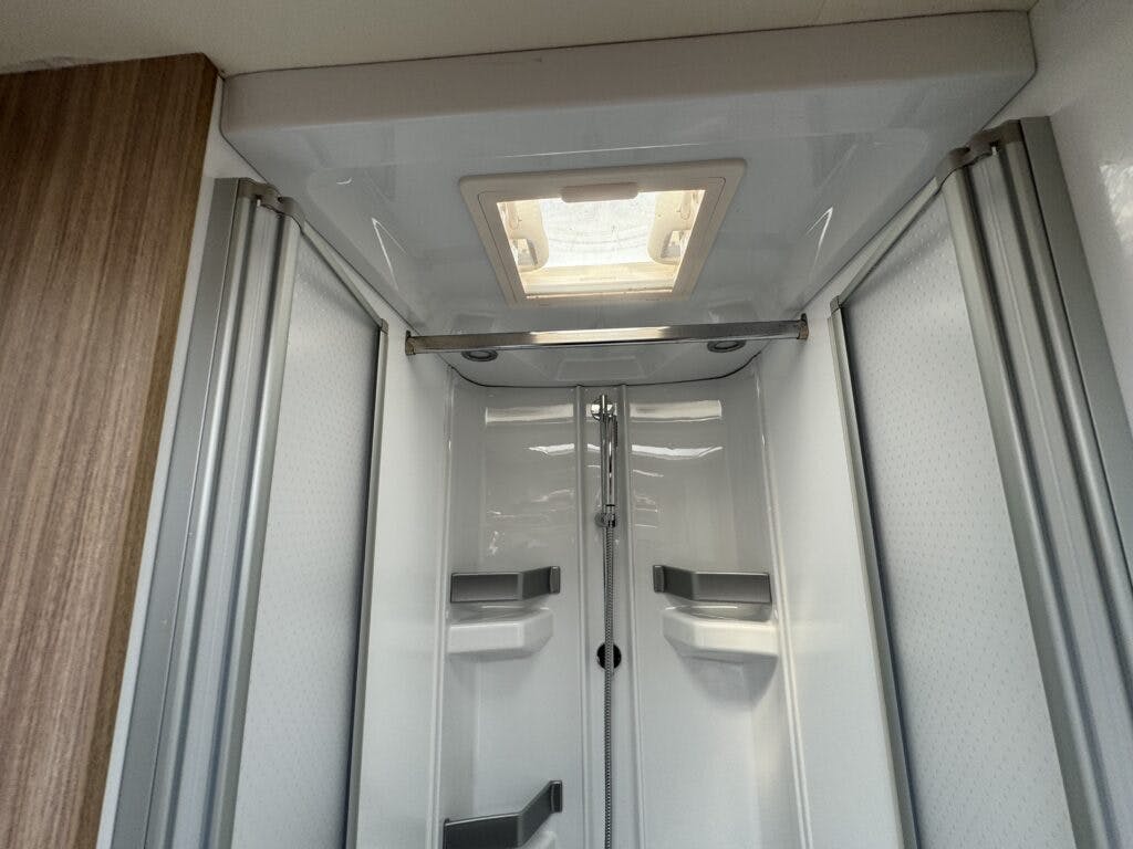 A compact shower cabin in the 2018 Burstner Ixeo TL680 G features white walls, a small overhead skylight, and a metal showerhead attached to the back wall. The enclosure boasts multiple built-in shelves and sliding glass doors with metal frames. A wooden panel is visible on the left.