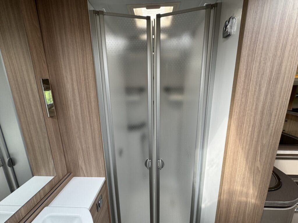 A narrow bathroom in the 2018 Burstner Ixeo TL680 G features wood paneling and a frosted glass shower enclosure with two folding doors. A white sink with a mirror above it is located to the left of the shower, while an unidentifiable room is visible in the background to the right.