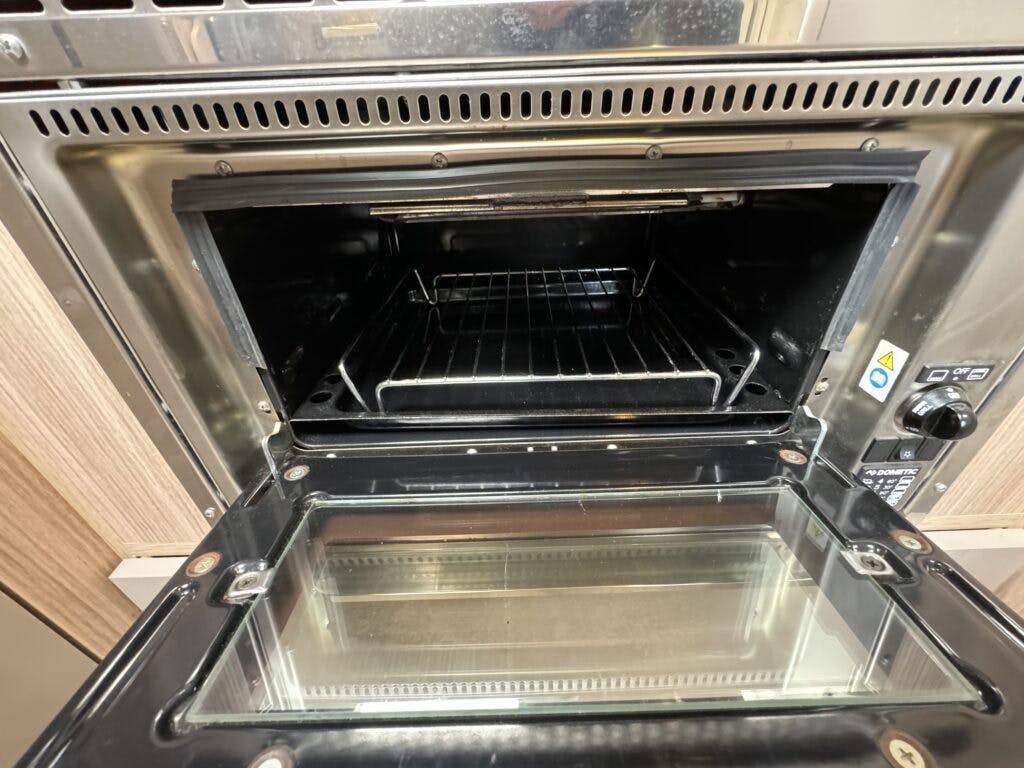 A photo of an open countertop oven in a 2018 Burstner Ixeo TL680 G. The glass door is open, revealing the interior with a wire rack. The oven controls are visible to the right, and the exterior is made of stainless steel. The surrounding cabinetry is light-colored.