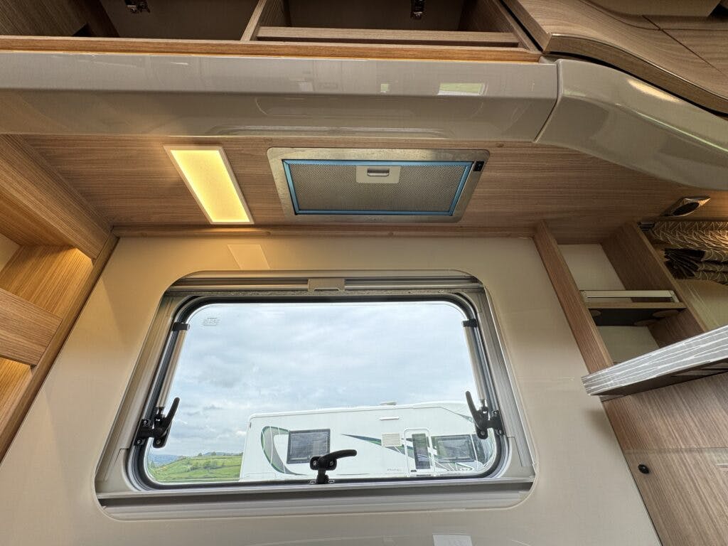 The interior roof and upper wall of a 2018 Burstner Ixeo TL680 G, showing a closed vent and a rectangular window. The window has black handles and looks out onto another caravan and a cloudy sky. Wooden cabinets are seen above the window.