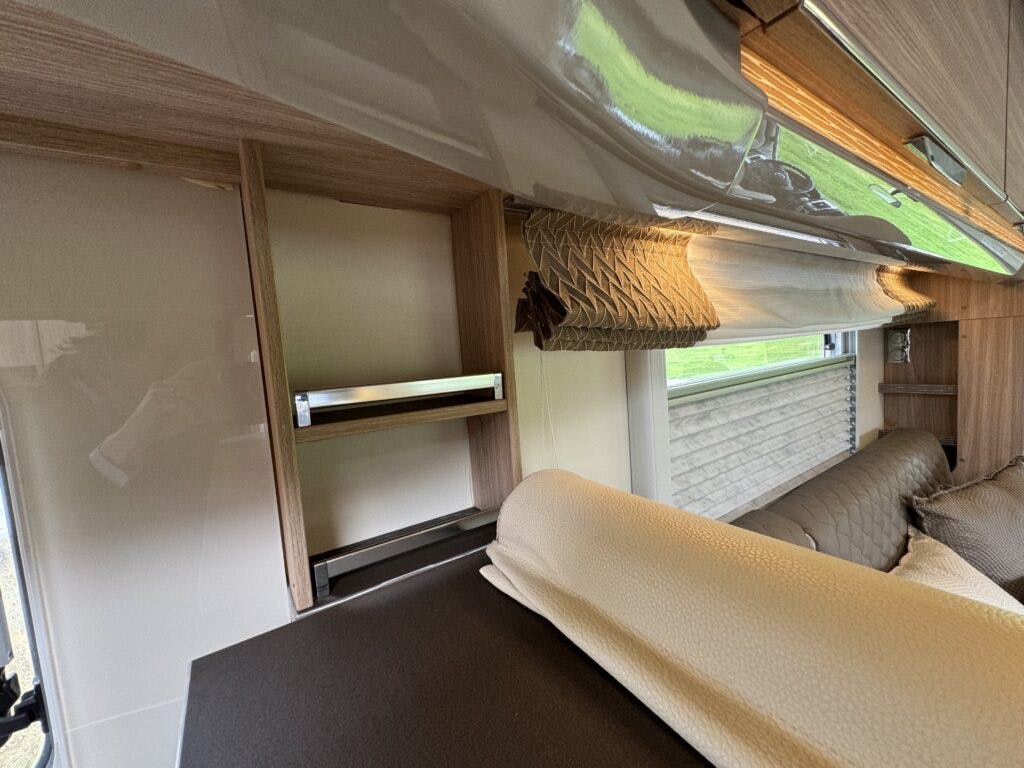 The image shows the interior of a 2018 Burstner Ixeo TL680 G camper van. It features a partially furnished storage compartment with a wooden frame, an empty shelf, a folded-down bed or seating area, and a window with a rolled-up blind. Green grass is visible outside the window.