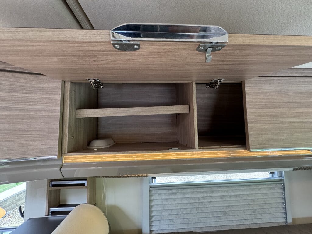 A view of an open wooden overhead cupboard inside a 2018 Burstner Ixeo TL680 G. The cupboard is divided into sections, with a wooden shelf and compartments. The ceiling and part of the interior, including a window with a blind, are visible in the background.
