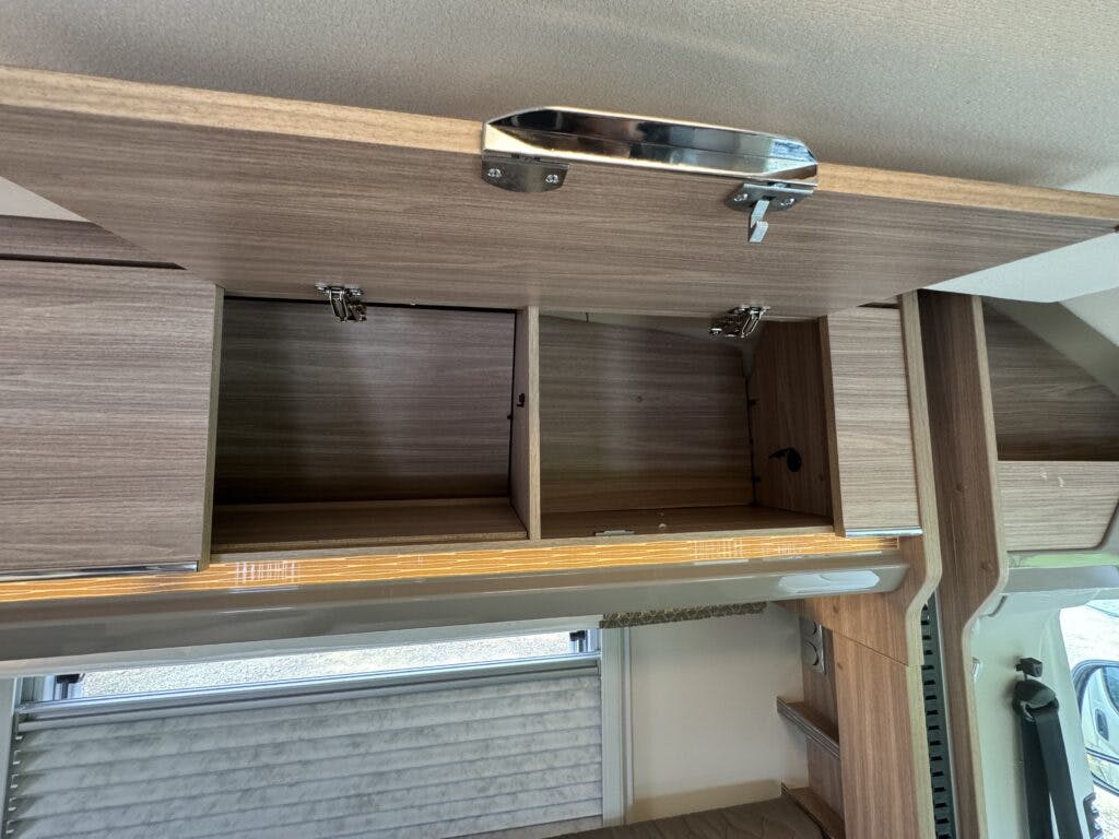 The image shows an open overhead storage cabinet inside a 2018 Burstner Ixeo TL680 G recreational vehicle (RV). The cabinet has several compartments, and the doors are hinged at the top. Below the cabinet, there is a window with a partially closed blind.