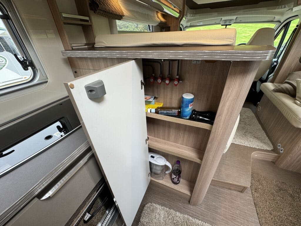 The interior of the 2018 Burstner Ixeo TL680 G camper van features an open cupboard door, revealing stored items including cleaning supplies, a water bottle, a paper towel roll, and various other small essentials. Above the cupboard is a bench seat, and to the left, you can see part of a compact kitchen area.