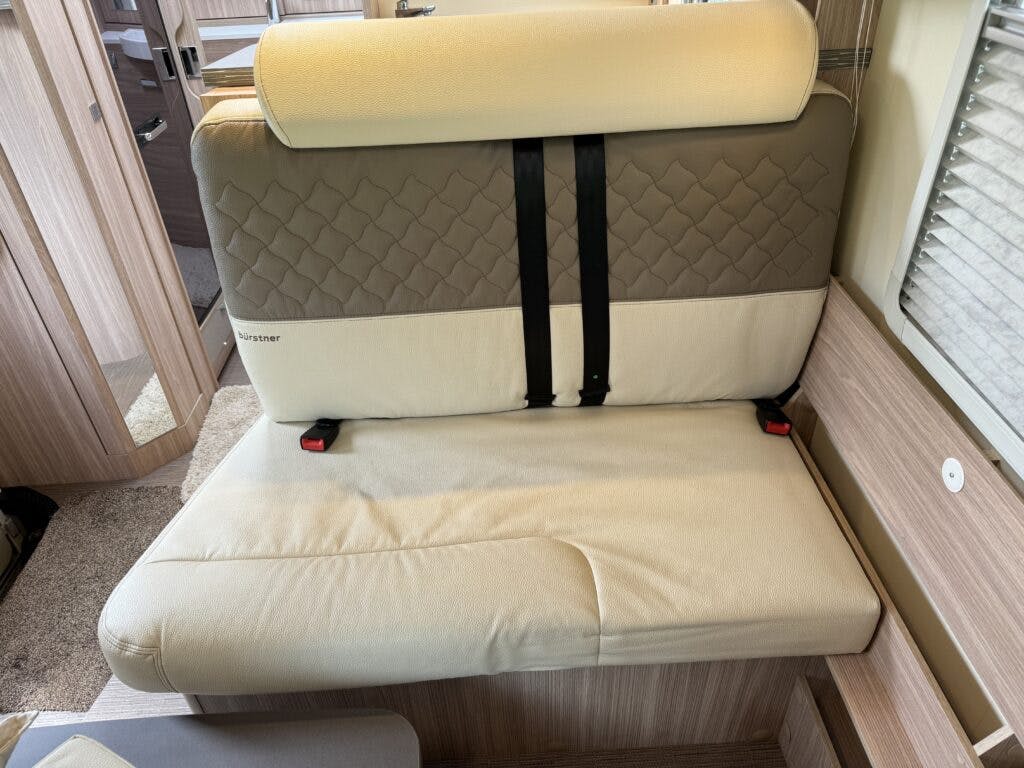 An upholstered bench seat with a padded backrest and seatbelt straps inside the 2018 Burstner Ixeo TL680 G camper. The seat features beige and light brown sections with a quilted pattern on the backrest, complemented by light wood paneling and a window on one side.