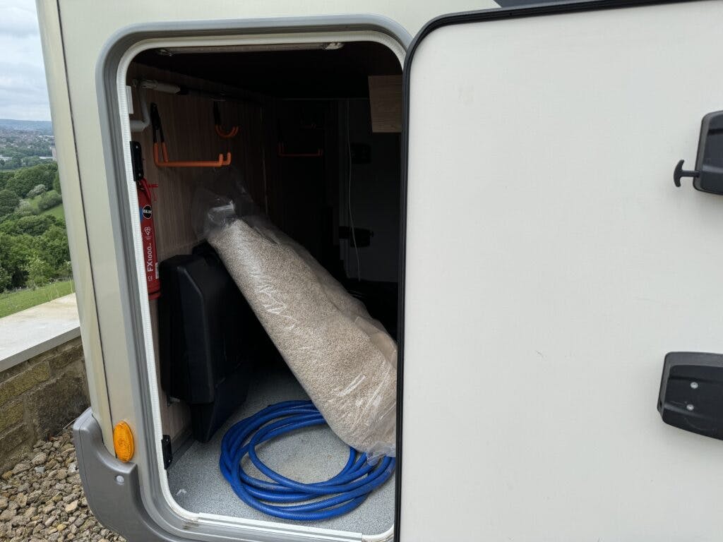 A storage compartment in the 2018 Burstner Ixeo TL680 G is shown, containing a rolled-up carpet, a blue hose, a black container, and a red fire extinguisher mounted on the side wall. The compartment door is open, with the scene set against a background of grass and shrubs.
