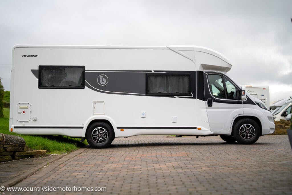 A white 2022 Benimar Mileo 282 motorhome is parked on a paved area beside a grassy surface. The vehicle features large tinted windows on the side and a black stripe with a logo. The sky is overcast, and a website URL is visible in the bottom-left corner of the image.