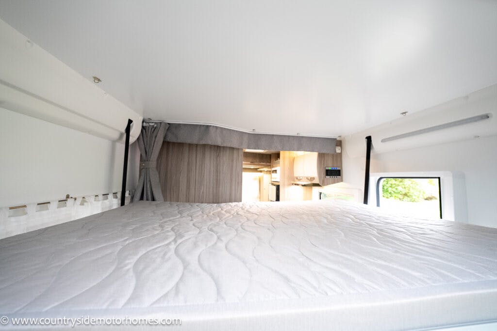 The image shows the interior of a 2022 Benimar Mileo 282 motorhome featuring a spacious, elevated sleeping area with a fitted mattress. There is a curtain that can be drawn for privacy. The background reveals a kitchen area with cabinets and appliances.