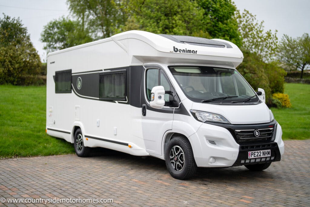 A white 2022 Benimar Mileo 282 motorhome is parked on a paved driveway. The vehicle boasts a clean, modern design with the Benimar logo on the front. It features a UK license plate with the registration PE22 NHH. In the background, there is a green lawn and trees.