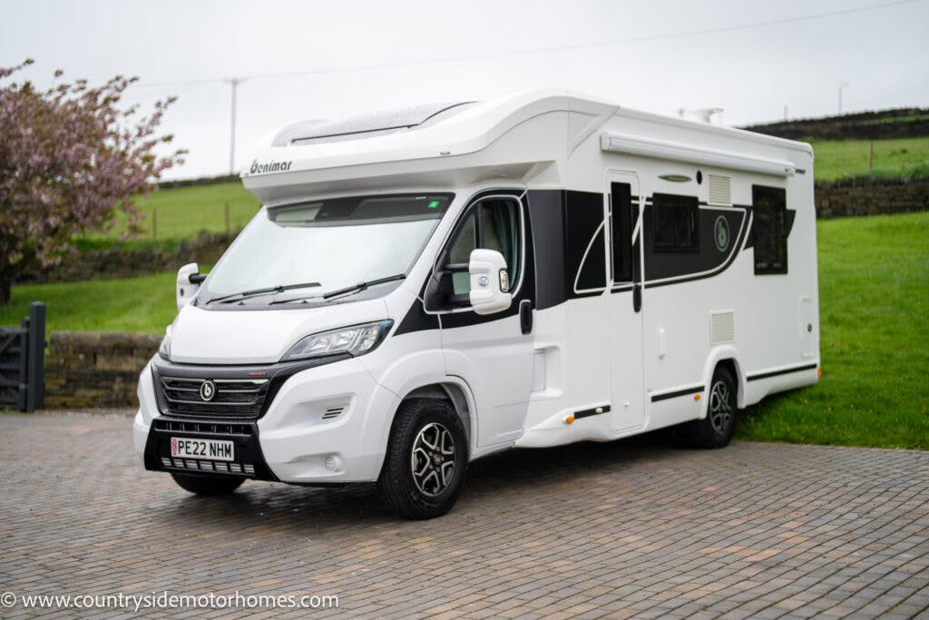 A 2022 Benimar Mileo 282 with the registration plate "PF22 HWH" is parked on a stone-paved driveway. The white motorhome features black and white detailing along the sides. In the background, there is grassy terrain, a stone fence, and several trees.