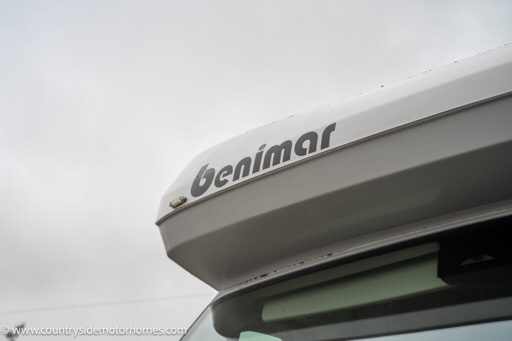 A close-up view of the upper front section of a 2022 Benimar Mileo 282 motorhome, featuring the brand name "Benimar" in black text on a white background. The sky is overcast. The vehicle is partly visible, showing the top of the windshield.