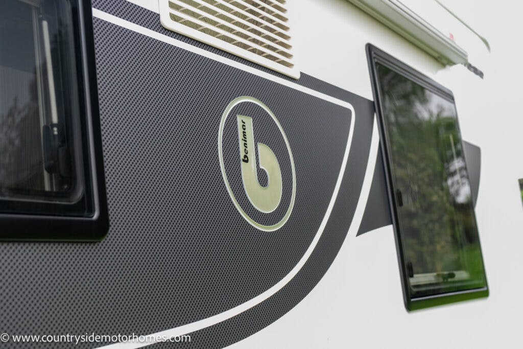 Close-up of a portion of a white motorhome exterior. Visible are a window, part of an air vent, and the "Benimar" brand logo on a grey patterned section. This 2022 Benimar Mileo 282 model showcases sleek design details. Photo credit: "www.countrysidemotorhomes.com" in the bottom left corner.