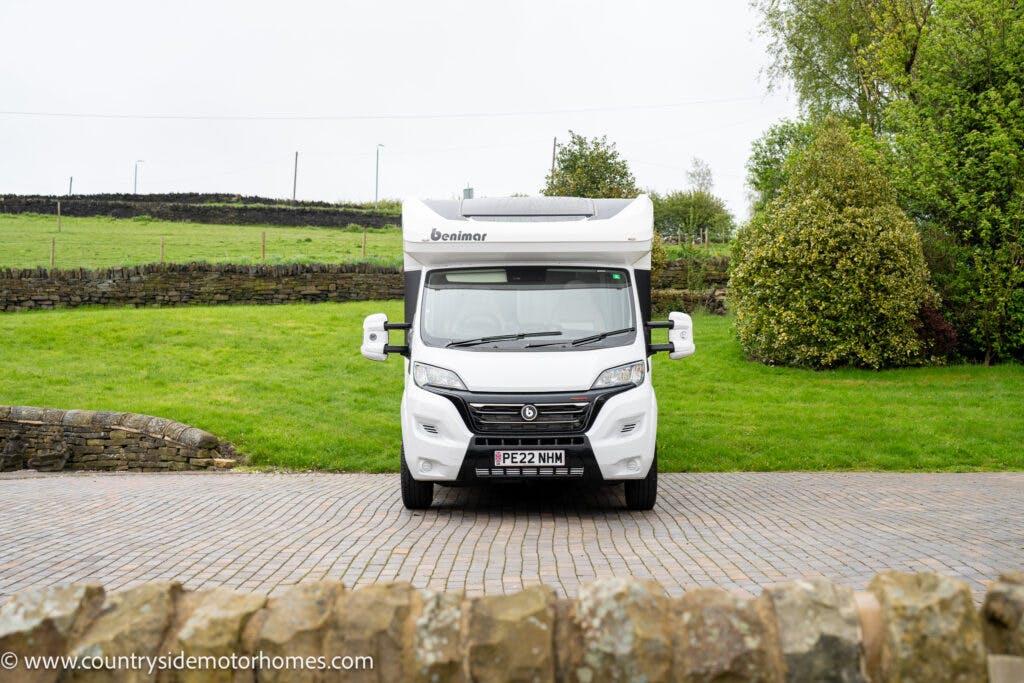 A white 2022 Benimar Mileo 282 motorhome is parked on a brick driveway with green fields and stone walls in the background. The vehicle has a registration plate reading "PE22 NWW." The surrounding area features various trees and bushes.