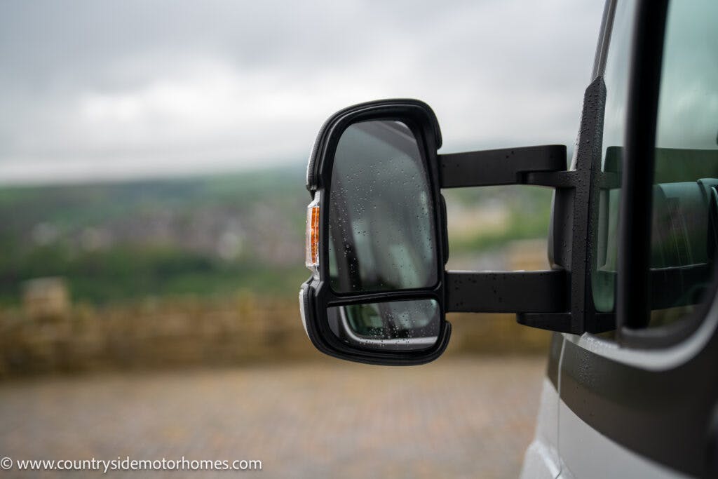 Close-up of a 2022 Benimar Mileo 282's side mirror on a rainy day. The mirror is wet with raindrops, and a blurred scenic background is visible. The website URL, www.countrysidemotorhomes.com, is seen in the bottom left corner of the image.