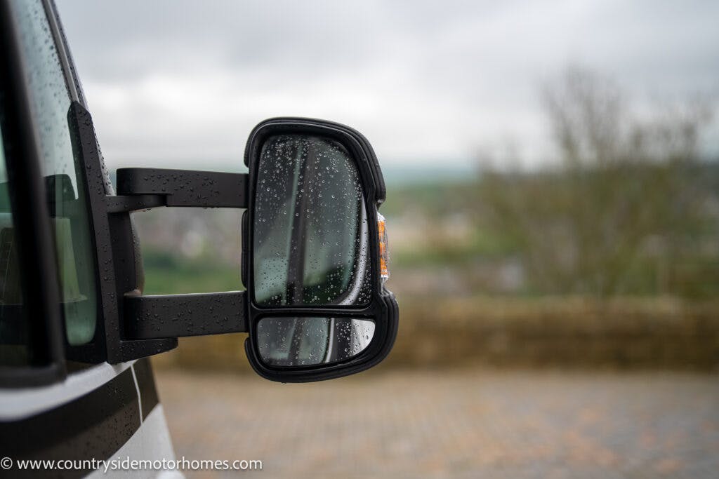 Close-up of a 2022 Benimar Mileo 282's side mirror on a rainy day. The mirror, wet with raindrops, reflects a blurred view of the surroundings. The background shows an out-of-focus landscape with overcast skies. A website URL is visible in the lower left corner.