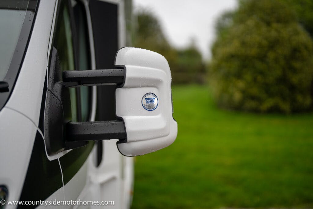 Close-up of a white side mirror on a 2022 Benimar Mileo 282 motorhome by Wing Mirror Guard. The mirror is slightly wet from rainfall. In the background, there is greenery, including some trees and bushes.