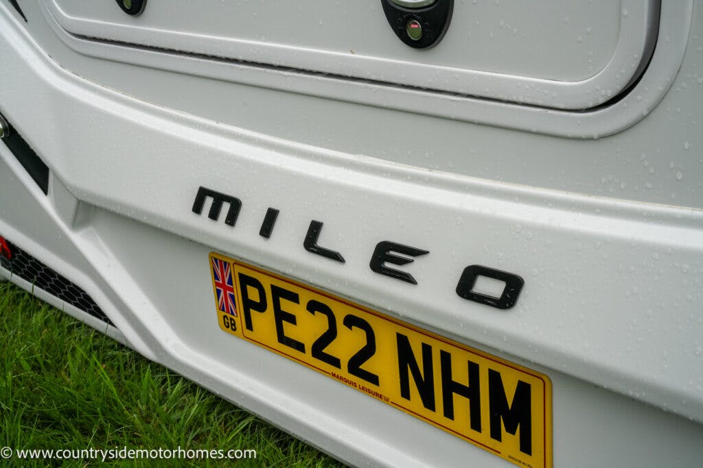 Close-up of the back of a 2022 Benimar Mileo 282 motorhome showing the "Mileo" logo and a yellow license plate with the registration "PE22 NHM." The motorhome is parked on grass on a rainy day, with water droplets visible on the surface. The website "countrysidemotorhomes.com" is in the bottom left corner.