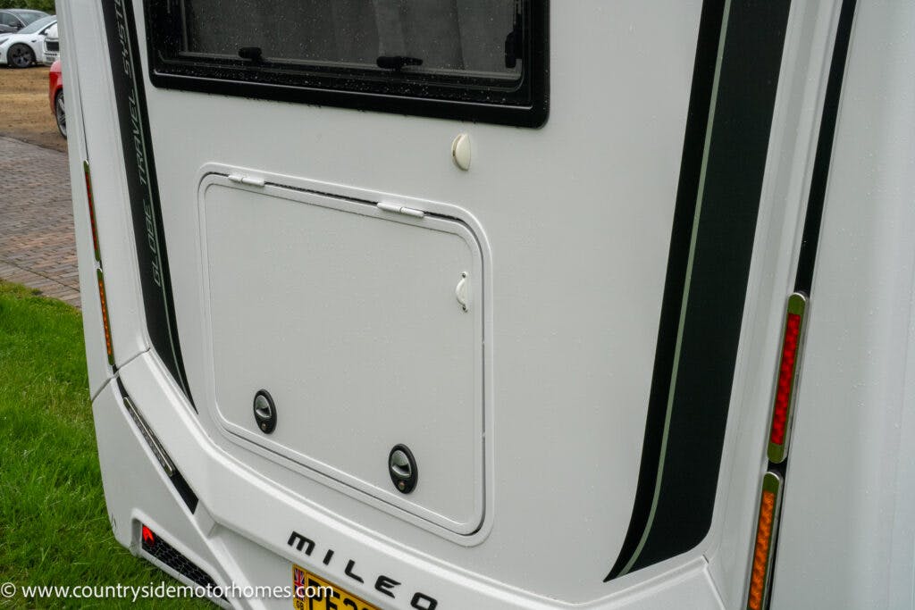 A close-up view of the rear of a 2022 Benimar Mileo 282 motorhome with the word "Mileo" visible. There are a few black accent lines and several red and amber reflectors. A panel with two round fasteners is present, and the background includes some grass and a paved area.