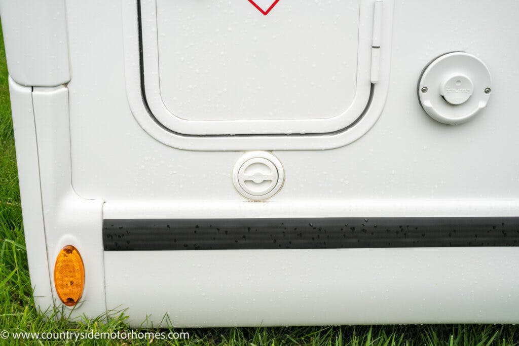 Close-up view of the side of a 2022 Benimar Mileo 282 motorhome, featuring various access panels and connections. The image shows a lockable door and circular fixtures, including a gas or water hookup and an indicator light near the bottom. Grass is visible below.