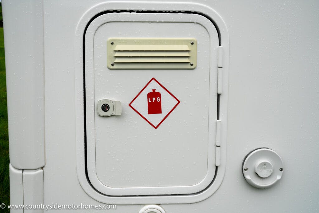 A white metal access panel on the 2022 Benimar Mileo 282 features a red diamond-shaped sign with a gas cylinder and the letters "LPG." The panel has a small vent above and a round latch below. To the right, there's a circular fixture. The surface appears to be wet.
