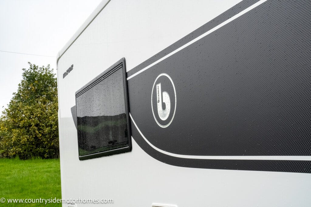 Close-up of the side of a 2022 Benimar Mileo 282 motorhome with black and grey detailing, featuring a small window and a logo. The surface appears wet from rain. The background includes a grassy area and trees. A website URL is visible at the bottom left corner: www.countrysidemotorhomes.com.