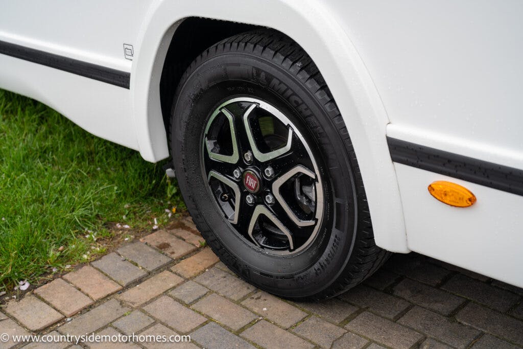 A close-up view of the 2022 Benimar Mileo 282 camper van wheel with a metallic black and silver rim. The van's white body is visible in the background, and the wheel is parked on a surface that transitions from grass to bricks. An amber side marker light is also visible.