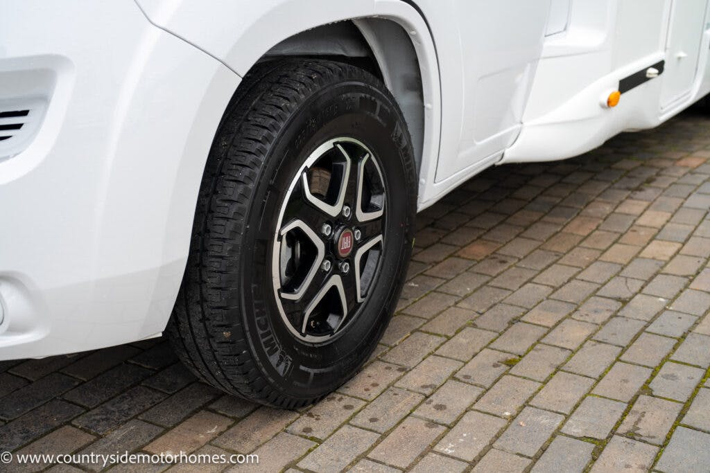 A close-up view of a black tire with a five-spoke alloy wheel on the 2022 Benimar Mileo 282. The vehicle is parked on a paved surface made of interlocking bricks. A website URL is visible in the bottom left corner of the image.