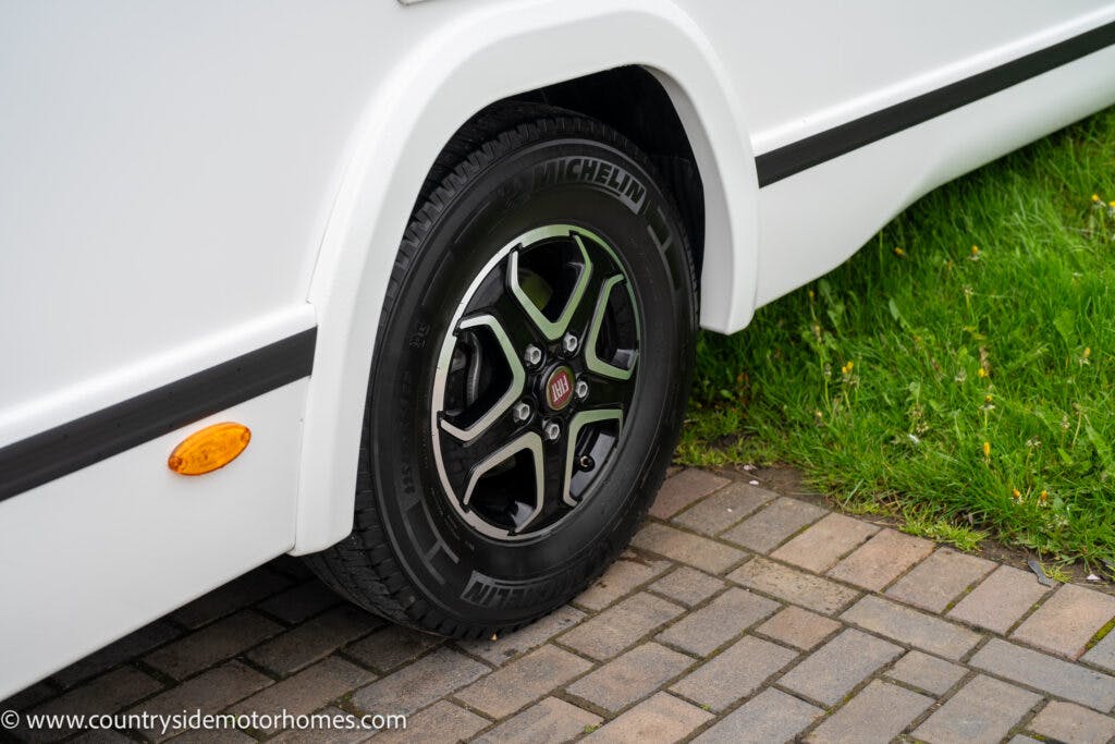 A close-up of a vehicle's wheel with a Michelin tire, featuring part grass and part brick surface. The wheel belongs to a 2022 Benimar Mileo 282, attached to a white body with a round orange reflector near the tire. The image's website URL is visible at the bottom-left corner.