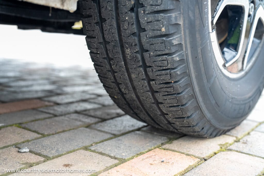Close-up image of a vehicle tire on a paved surface. The pavement consists of rectangular, interlocking bricks in varying shades of gray and brown. The tire tread appears worn and has some dirt on it. Just beyond the tire, "www.countrysidemotorhomes.com" advertises the 2022 Benimar Mileo 282.