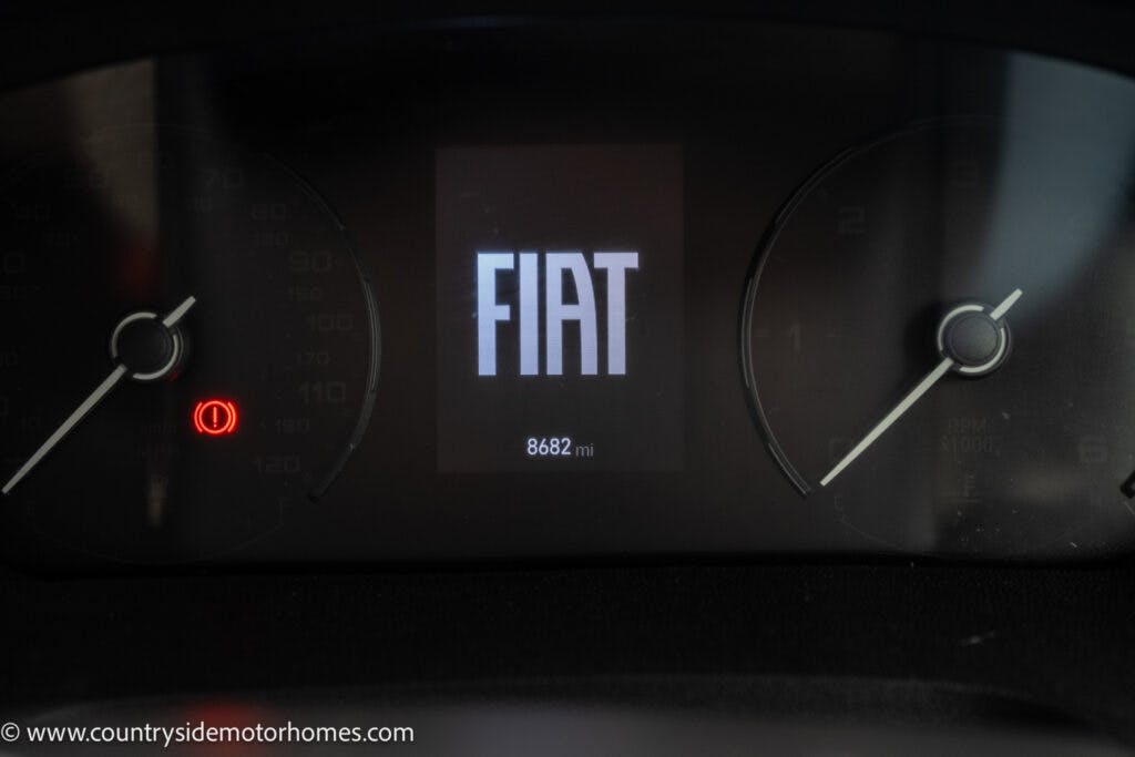 A close-up view of a car dashboard showing the Fiat logo in the center display. The odometer reads 8682 miles. The dark dashboard background features two circular gauges on either side, similar to those found in a 2022 Benimar Mileo 282. A small red indicator light is illuminated on the left gauge.