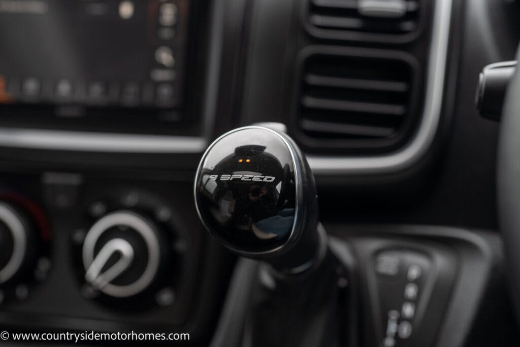 Close-up of a 6-speed manual gear shift knob inside a 2022 Benimar Mileo 282. The background shows part of the car's dashboard, including air vents and climate control knobs. The text "6 SPEED" is visible on the gear shift knob.