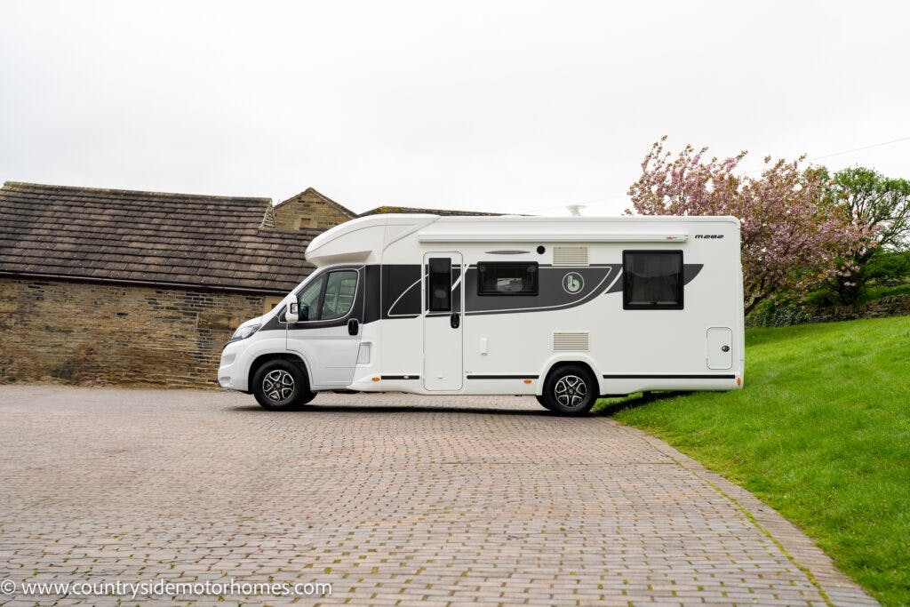 A white 2022 Benimar Mileo 282 motorhome with black decals is parked on a paved driveway next to a grassy area and a stone building. There are blooming trees in the background. The web address www.countrysidemotorhomes.com is visible in the bottom-left corner of the image.