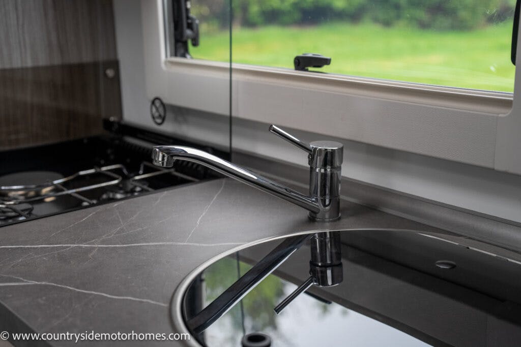 Image of a modern kitchen sink area in the 2022 Benimar Mileo 282 motorhome. The sink has a sleek, chrome faucet and is set into a dark countertop with a marble-like pattern. There is a window above the sink looking out to a green, blurred outdoor landscape.
