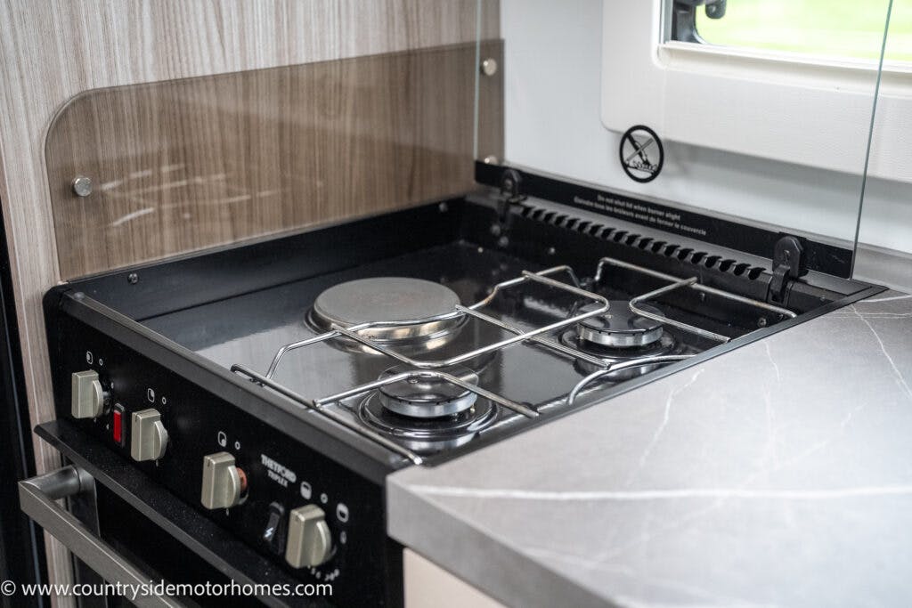 A close-up view of a two-burner gas stove in the stylish kitchen of the 2022 Benimar Mileo 282. The stove features two control knobs below the burners, with a window visible in the background. The surrounding surfaces are elegantly covered in light wood and grey countertop materials.