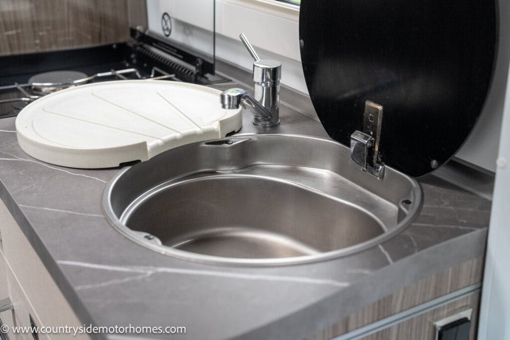 A compact kitchen sink with a closed soap dispenser and faucet, adjacent to a partially open stovetop cover, in a modern 2022 Benimar Mileo 282 motorhome. The countertop is a dark marble-like material, and the background shows part of a stove.