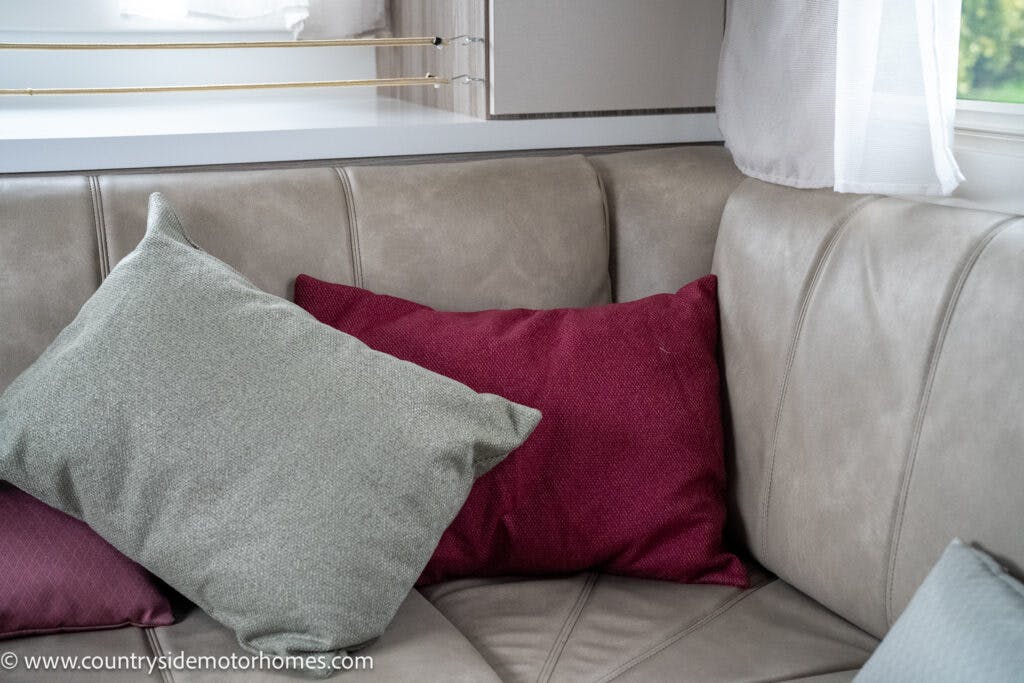 A corner of a taupe-colored leather sofa with three cushions placed on it. Two cushions are red, and one is green. White curtains are partially visible in the background, and a cabinet from the 2022 Benimar Mileo 282 is mounted on the wall above the sofa.