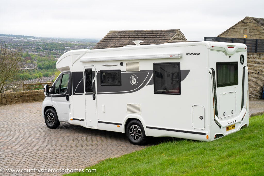 A modern white 2022 Benimar Mileo 282 motorhome is parked on a paved driveway, with a grassy area and brick building in the background. The motorhome features large windows and a side awning, set against a slightly overcast sky.