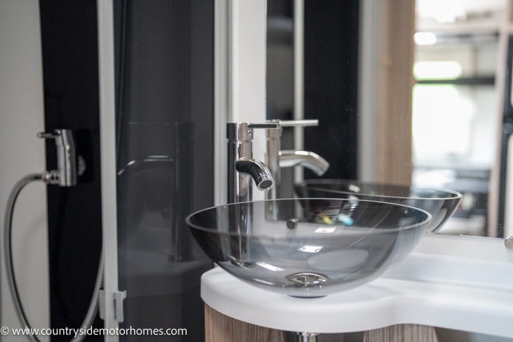 A modern bathroom sink in the 2022 Benimar Mileo 282 features a transparent glass bowl and a sleek, metallic faucet. There is a mirrored background reflecting the sink, and a partial view of a handheld showerhead can be seen on the left. The space appears clean and minimalist.