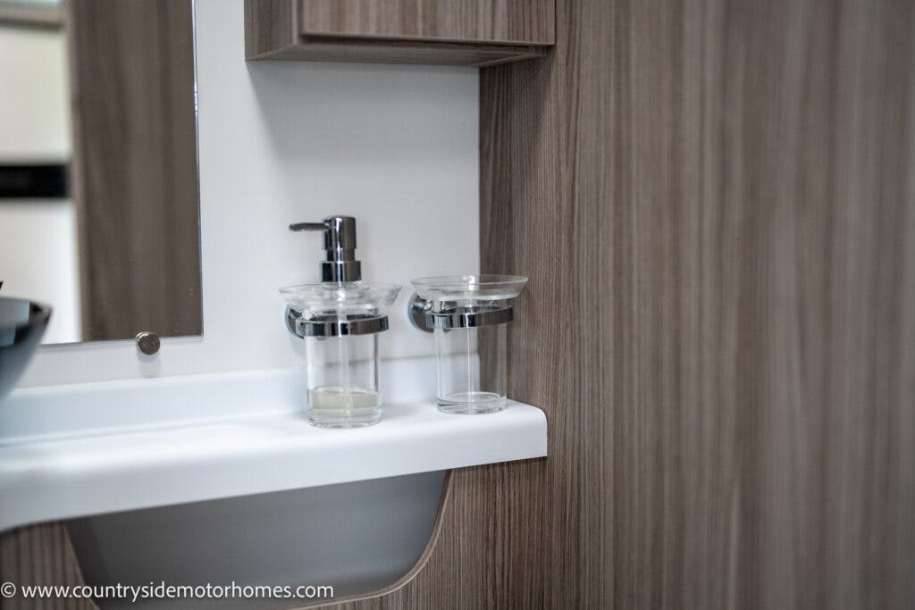 A modern bathroom sink area in the 2022 Benimar Mileo 282 features a mounted soap dispenser and a clear glass on a white surface, complemented by wooden paneling on the walls. A small mirror and part of a vanity cabinet are visible. The URL "www.countrysidemotorhomes.com" is at the bottom-left corner.