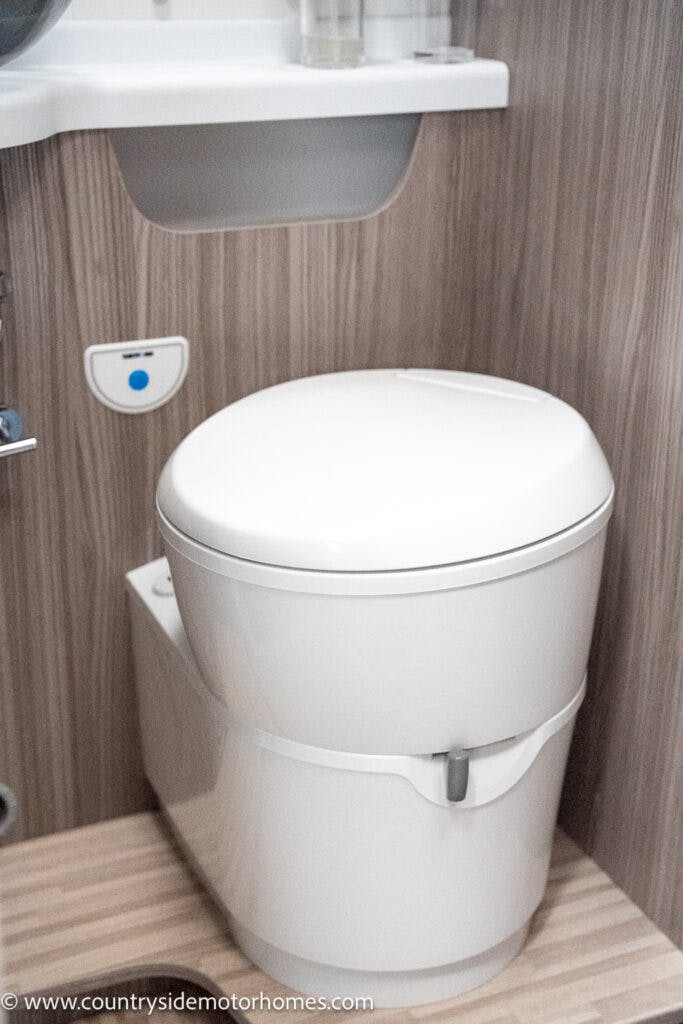 A compact white toilet with a curved lid is installed in a small bathroom with wooden paneling inside the 2022 Benimar Mileo 282. A sink with an under-shelf holding toiletries is visible above, while the floor features a light wood design. The image is watermarked with "www.countrysidemotorhomes.com.