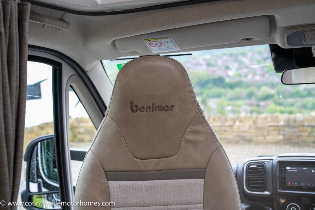 Interior view of the 2022 Benimar Mileo 282 motorhome focusing on the driver's seat. The seat is upholstered in light brown and beige fabric with "Benimar" stitched into the headrest. The dashboard, rearview mirror, and side window are partially visible. There is a scenic view outside.