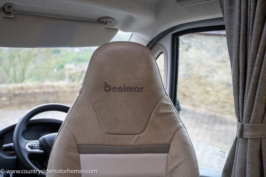 The image shows the interior of a 2022 Benimar Mileo 282 motorhome, specifically the driver's seat and window area. The seat has "Benimar" embroidered on the headrest. The steering wheel and part of the dashboard are visible, while the window curtain is partially drawn.