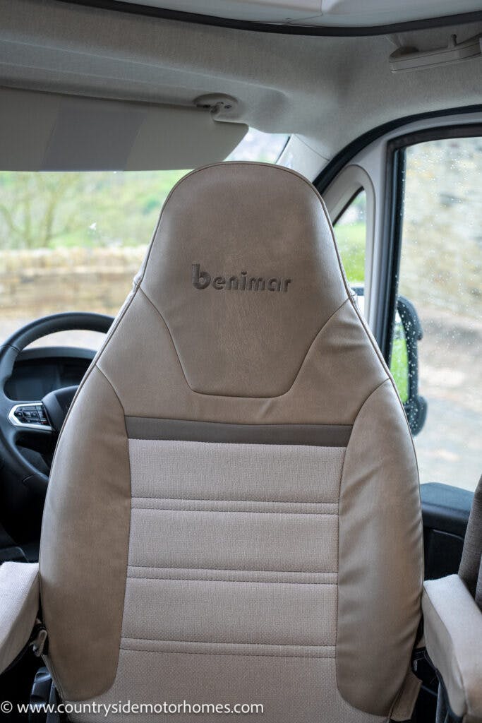 Front seats of a 2022 Benimar Mileo 282 motorhome with the brand name "Benimar" embroidered on the headrest. The upholstery is a combination of light brown and beige, with a sleek, modern design. The steering wheel and part of the dashboard are visible in the background.