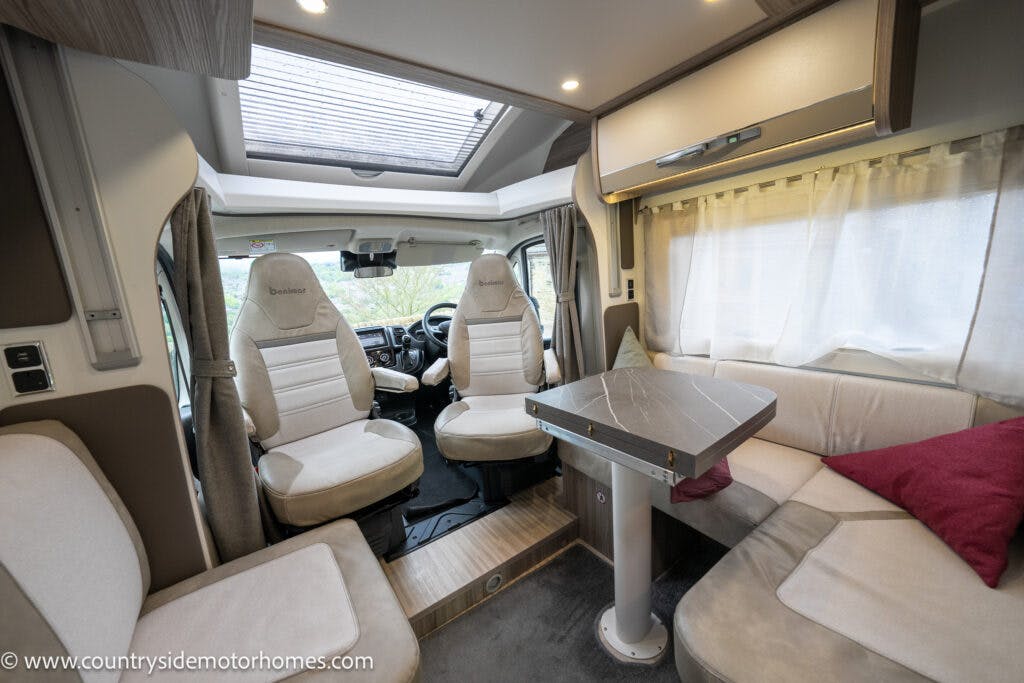 Interior view of the 2022 Benimar Mileo 282 RV with two front seats, a small table, and a cushioned seating area around it. The space has beige upholstery, a roof window, and a curtain covering a side window. The flooring is partially carpeted, and a red pillow is on the cushioned seating.