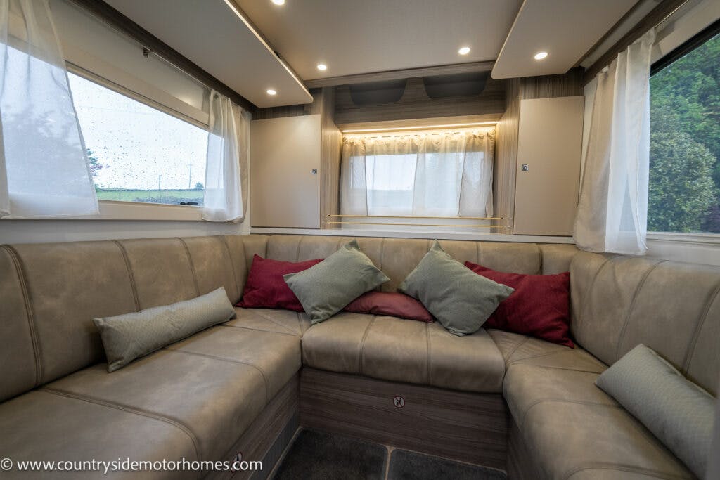 A modern motorhome interior of the 2022 Benimar Mileo 282 features a U-shaped beige leather seating area adorned with multiple throw pillows in green, red, and beige. Large windows with white curtains surround the seating, while wood-patterned flooring adds warmth. Text: www.countrysidemotorhomes.com in the lower left corner.