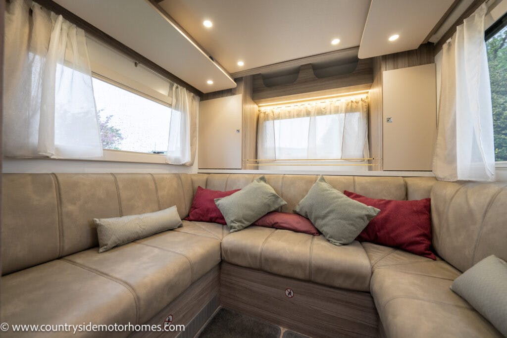 The interior of the 2022 Benimar Mileo 282 motorhome features a beige U-shaped sofa adorned with various colored throw pillows. The space is well-lit from windows on either side, and there are wooden cabinets above the sofa.